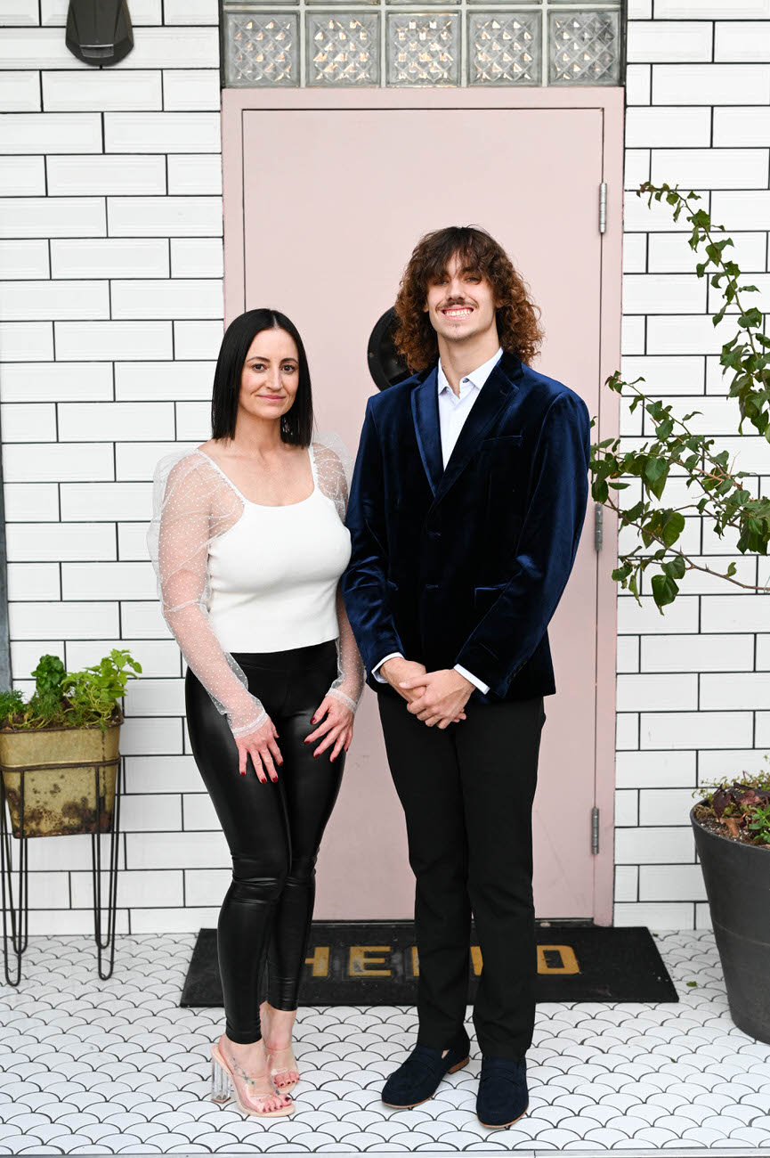 A mother and her adult son, dressed smartly, stand in front of a white brick wall by a pink door; the son in a blue velvet jacket and the mother in a white top with sheer sleeves and black pants.