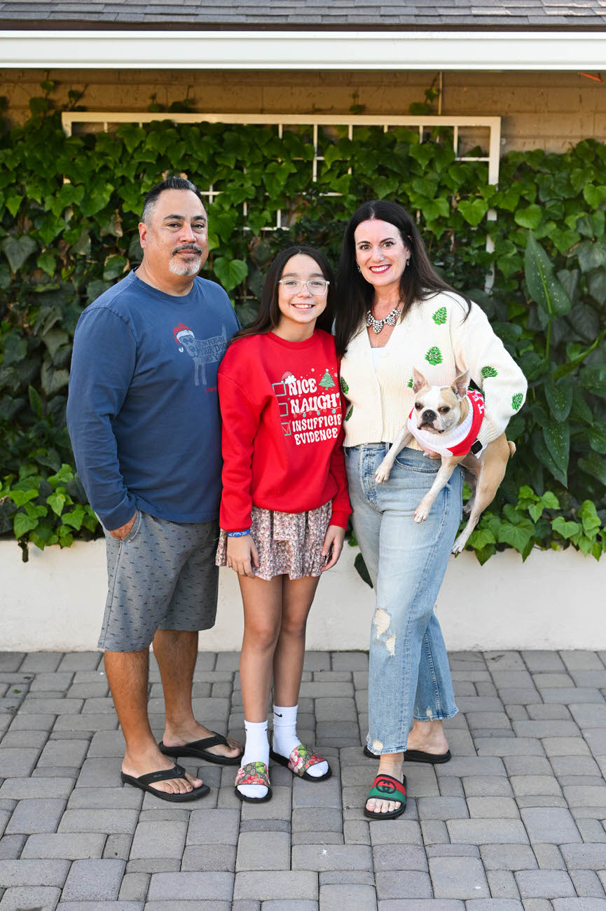 A family of three with their pet dog, smiling in front of a green vine-covered wall; the mother in a festive sweater and the daughter in a red "Naughty" Christmas sweater.