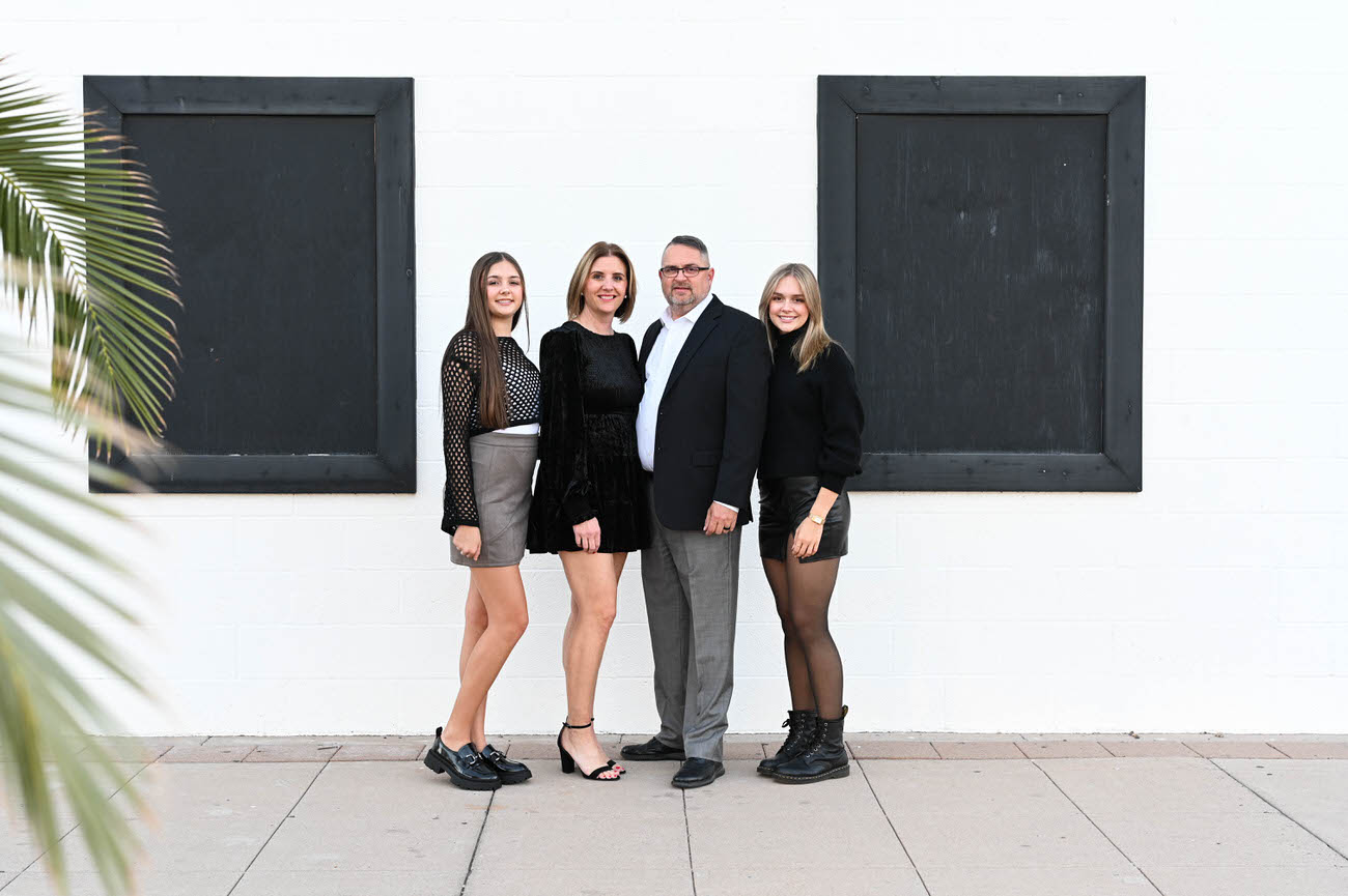 A family of four dressed in semi-formal attire, posing in front of a white wall with black shutters; the daughters in fashionable black outfits and the parents in coordinated black and grey.