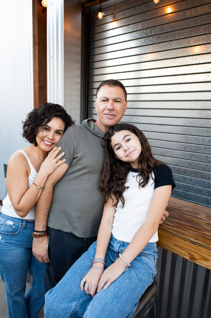 A family of three leaning against a wooden bar, the father in a grey hoodie, the mother in a white tank top, and the daughter in a black t-shirt, all in casual denim.