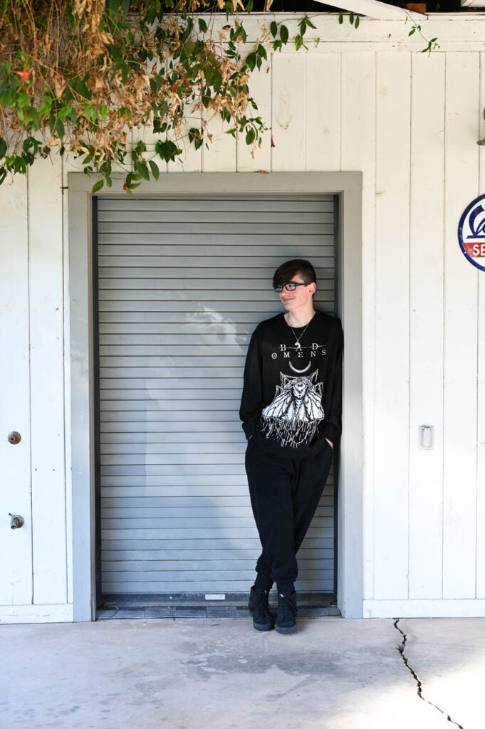 A young person in a black graphic hoodie and sunglasses stands alone, leaning against a grey roller shutter door, looking off to the side.