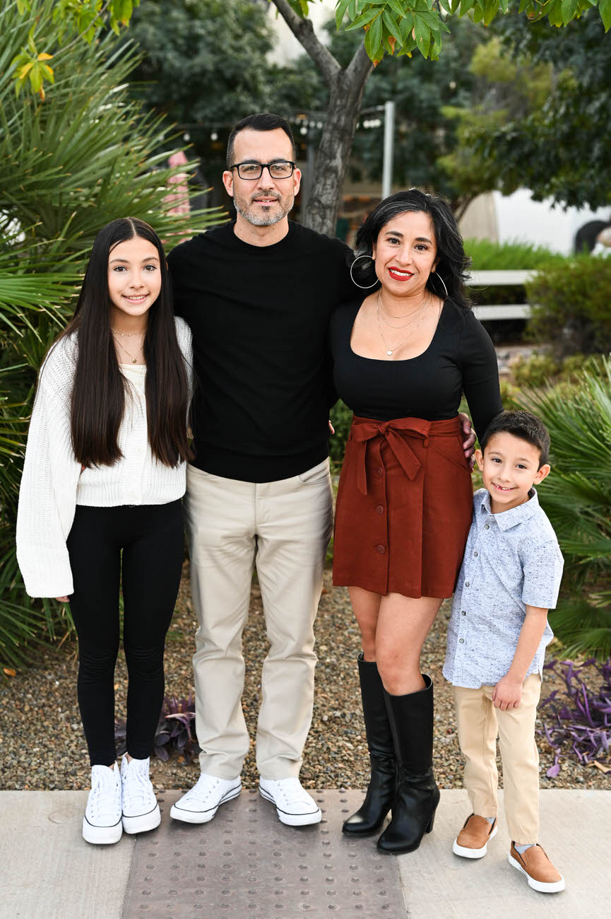 A family of four standing on a textured walkway; the daughters in white tops and black bottoms, the father in black and beige, and the mother in a black top and brown skirt with tall boots.