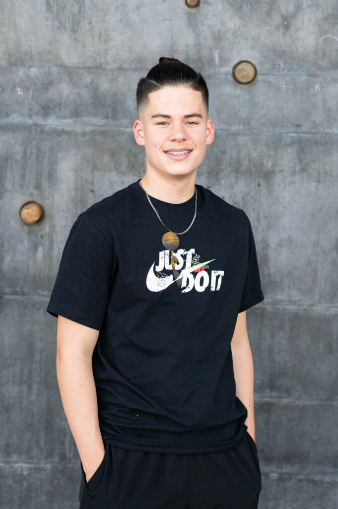 A young man in a casual black "Just Do It" t-shirt and shorts stands against a textured concrete wall, smiling at the camera.