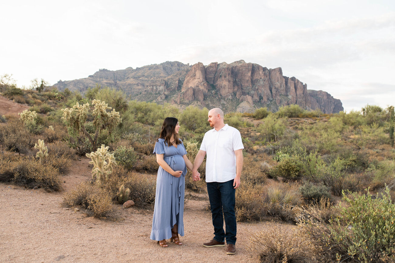 Expectant couple holding hands and gazing at each other, standing on a desert trail with tall cacti and a mountainous backdrop.