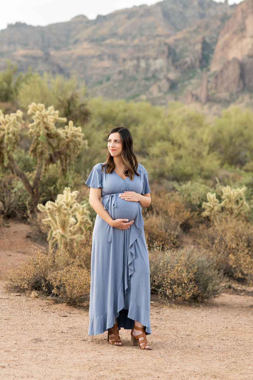 A pregnant woman in a flowing blue dress cradling her bump, standing alone on a desert path, looking to the side with a serene expression.