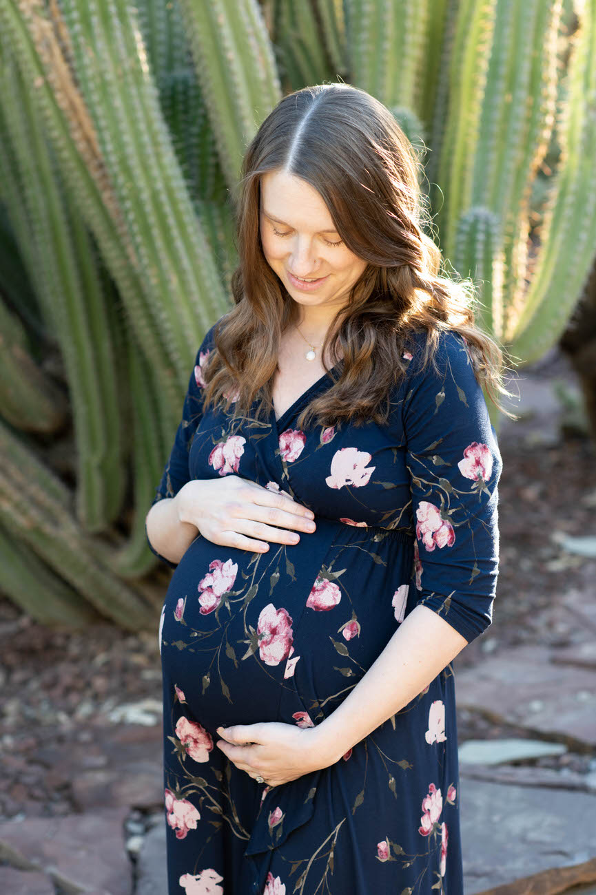 A side profile of a smiling pregnant woman in a floral dress, tenderly holding her bump with a backdrop of tall cacti in soft sunlight.