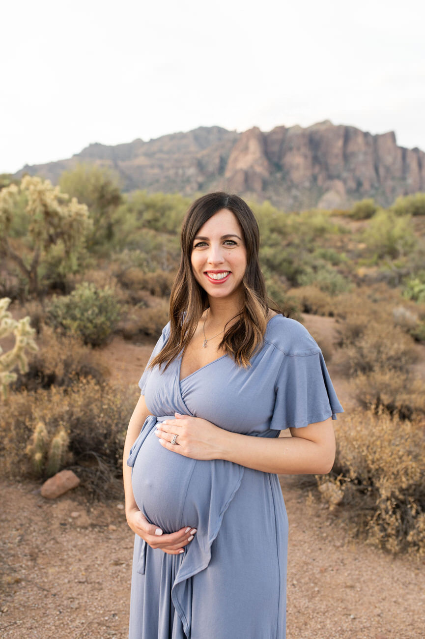 A straight-on portrait of a happy pregnant woman in a blue dress holding her belly, with a desert landscape in the background.