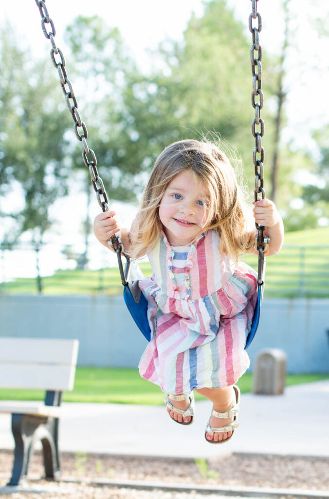 A young girl with long blonde hair enjoying a swing in a park, with a joyful expression on a sunny day.