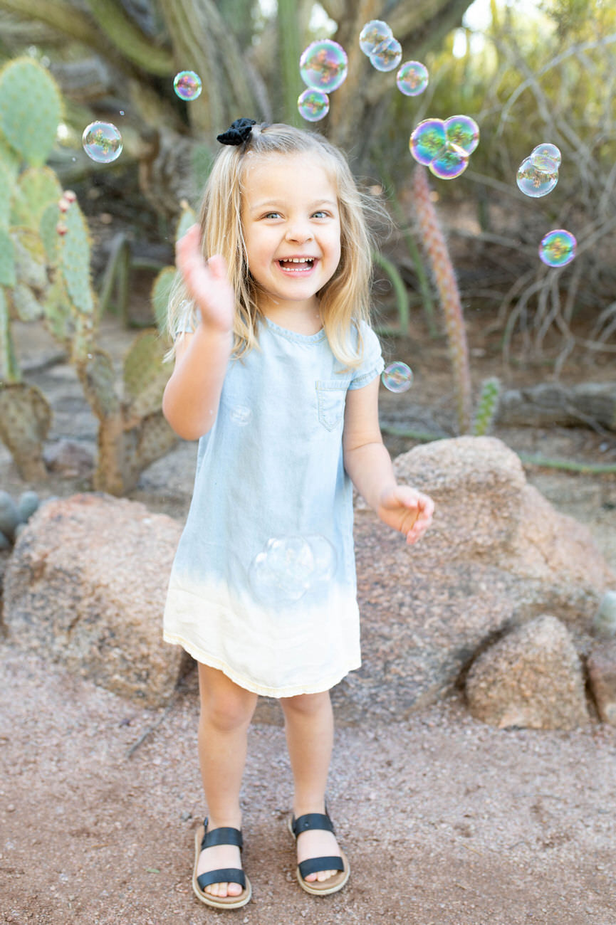 A joyful young girl with blonde hair wearing a blue dress and sandals playing with soap bubbles in a desert garden setting.