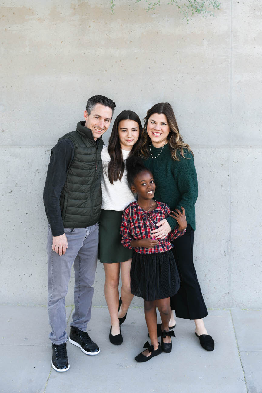 A family of four, including a teen girl, a young adopted girl, and their parents, posing with smiles in front of a concrete wall.
