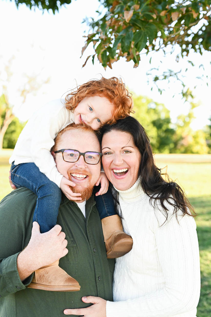 A portrait of a family of three with a redhead child sitting on her father's shoulders, all sharing a laugh against a backdrop of greenery.