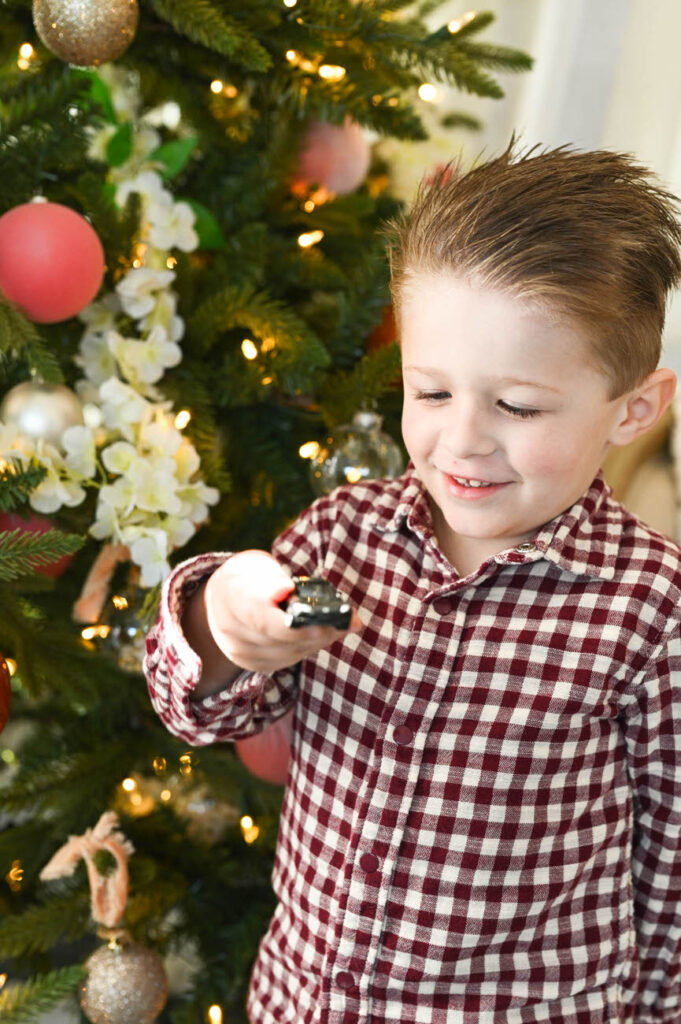 A little boy in a checkered shirt holding a toy car with a delighted expression by a Christmas tree.
