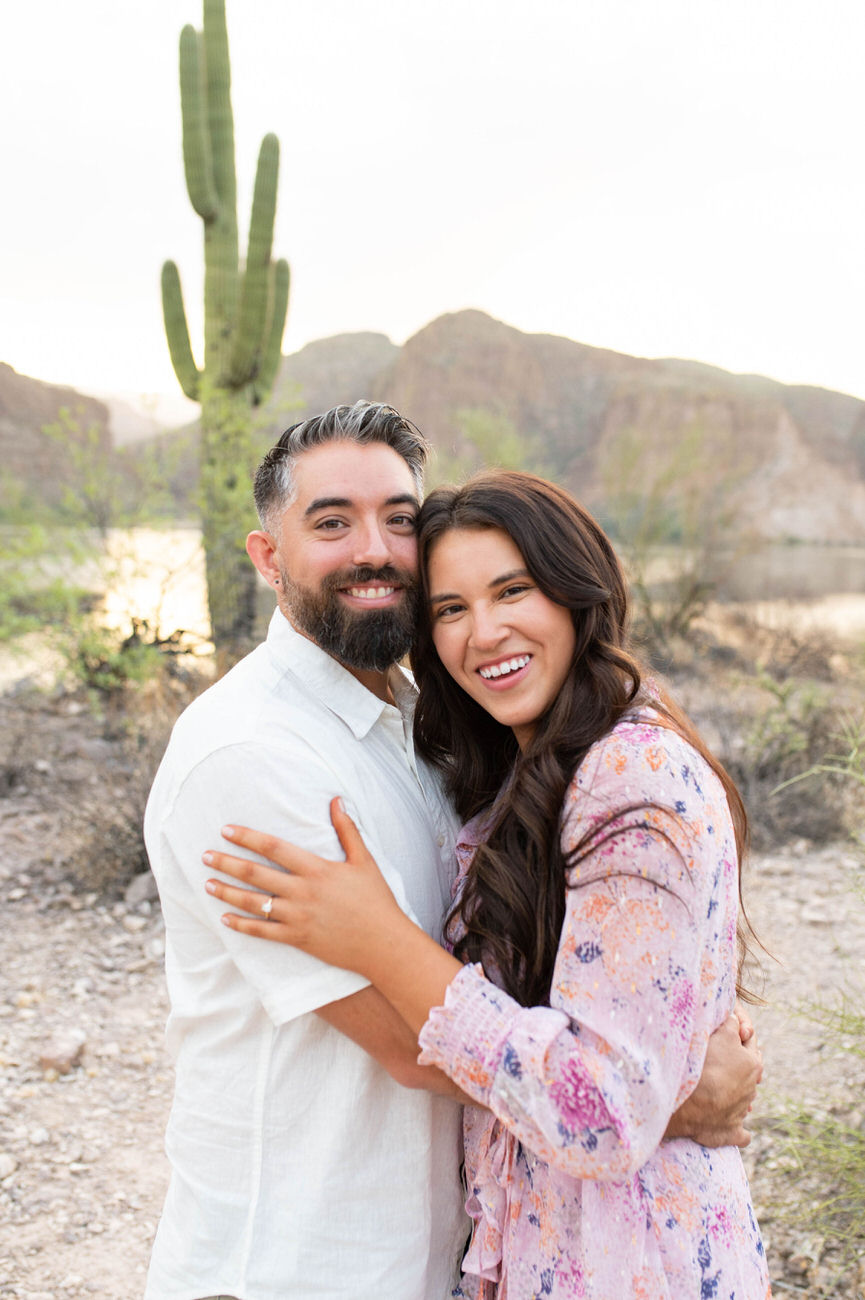 A couple embracing and smiling in a desert scene with a tall cactus in the background, conveying warmth and affection.