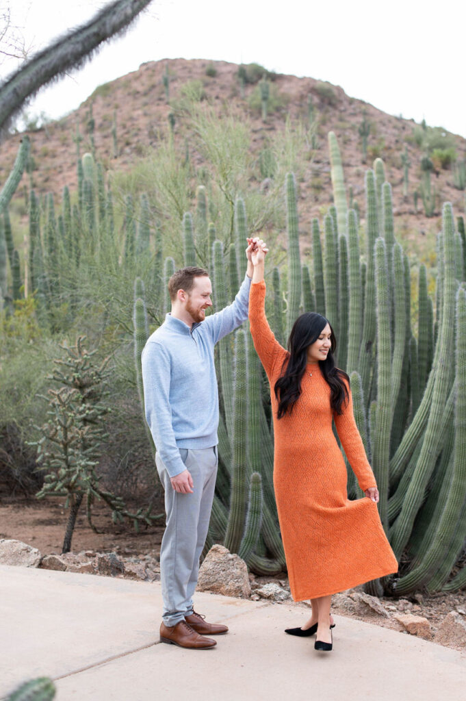 A couple dancing in a desert garden with a woman in an orange dress leading the man in a playful twirl, illustrating joy and companionship.