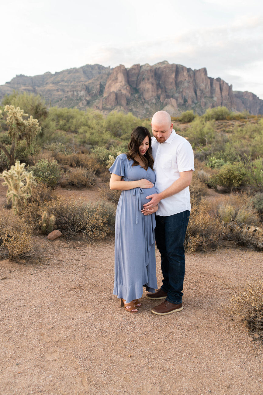 n expecting couple holding each other and the woman's belly, standing in a desert landscape, symbolizing anticipation and love.