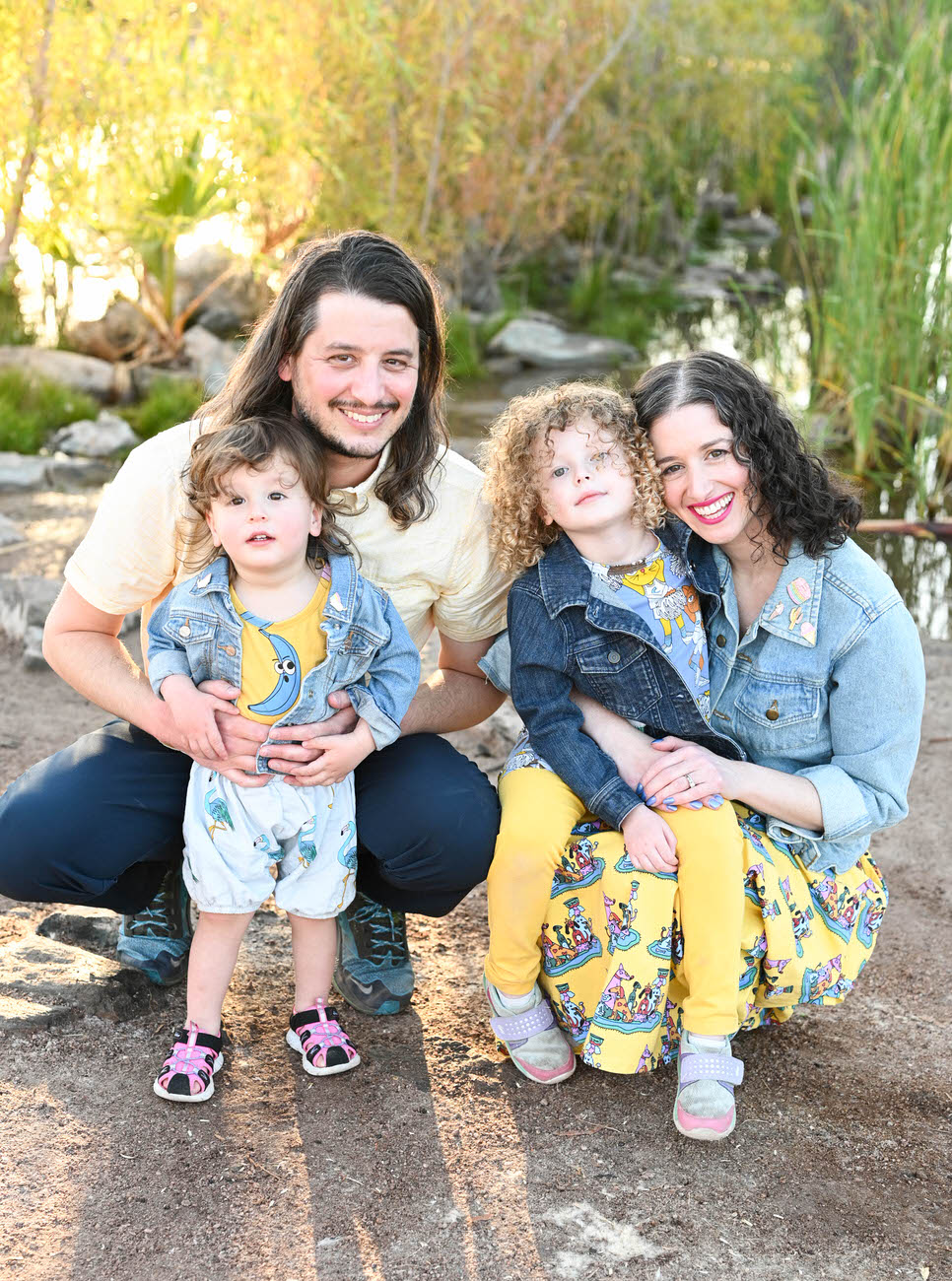 A joyful family of four with two young children wearing denim and yellow, posing together in an outdoor setting with lush greenery in the background.