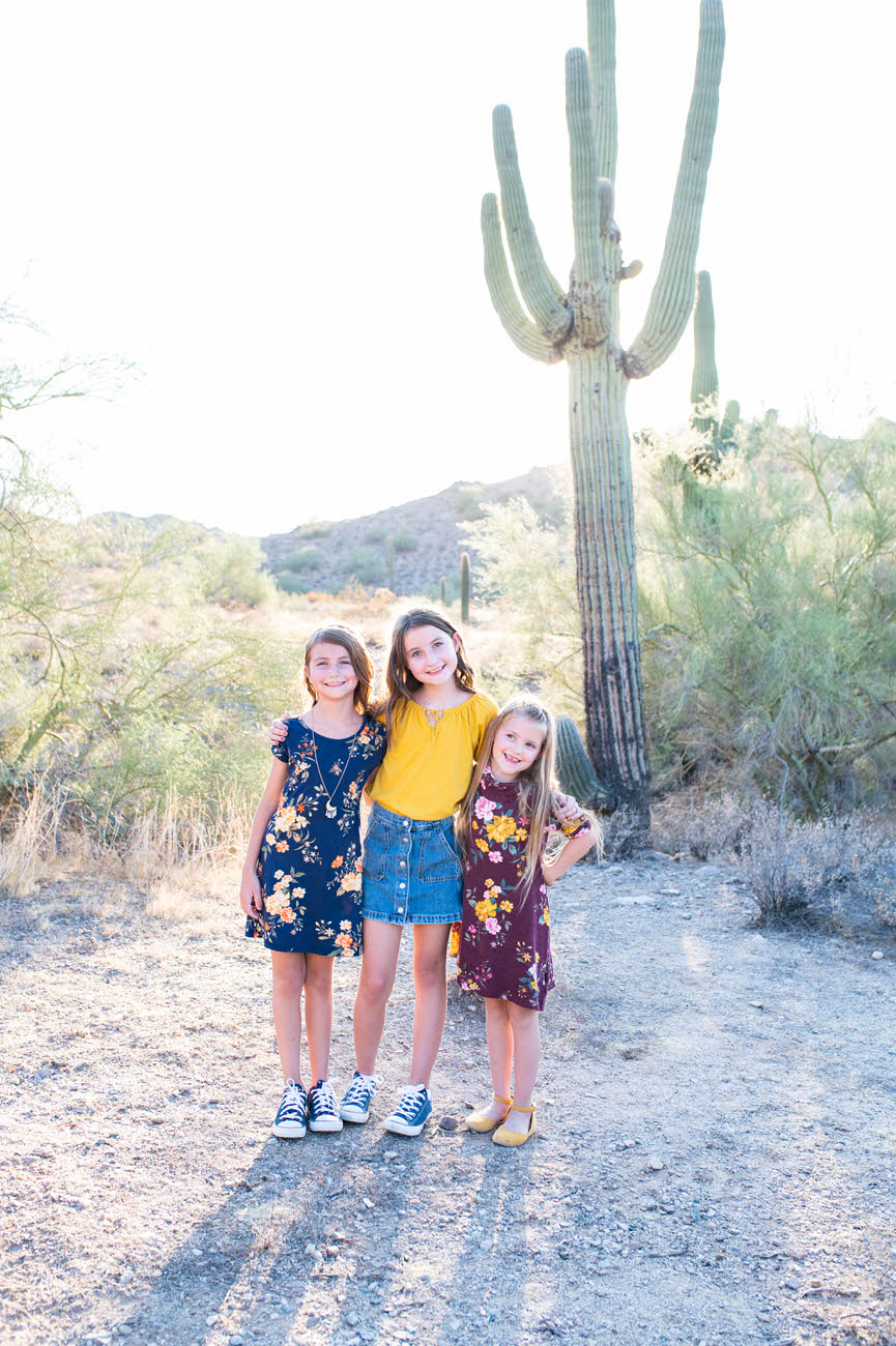 Three smiling young girls standing on a desert trail with a large saguaro cactus in the background, showcasing a sunny and warm environment.