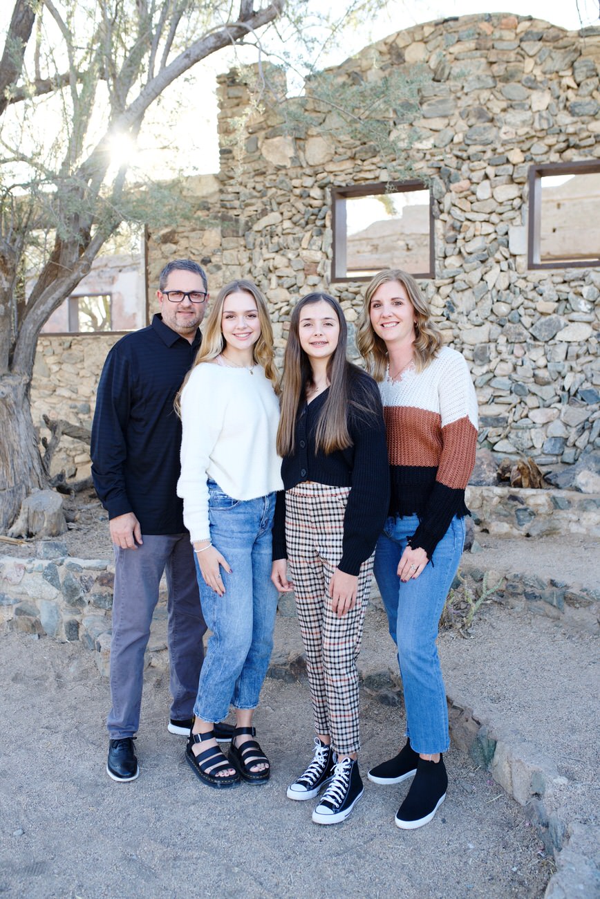 A family of four, with two teenage girls, dressed in smart casual attire posing in front of a rustic stone building surrounded by trees.