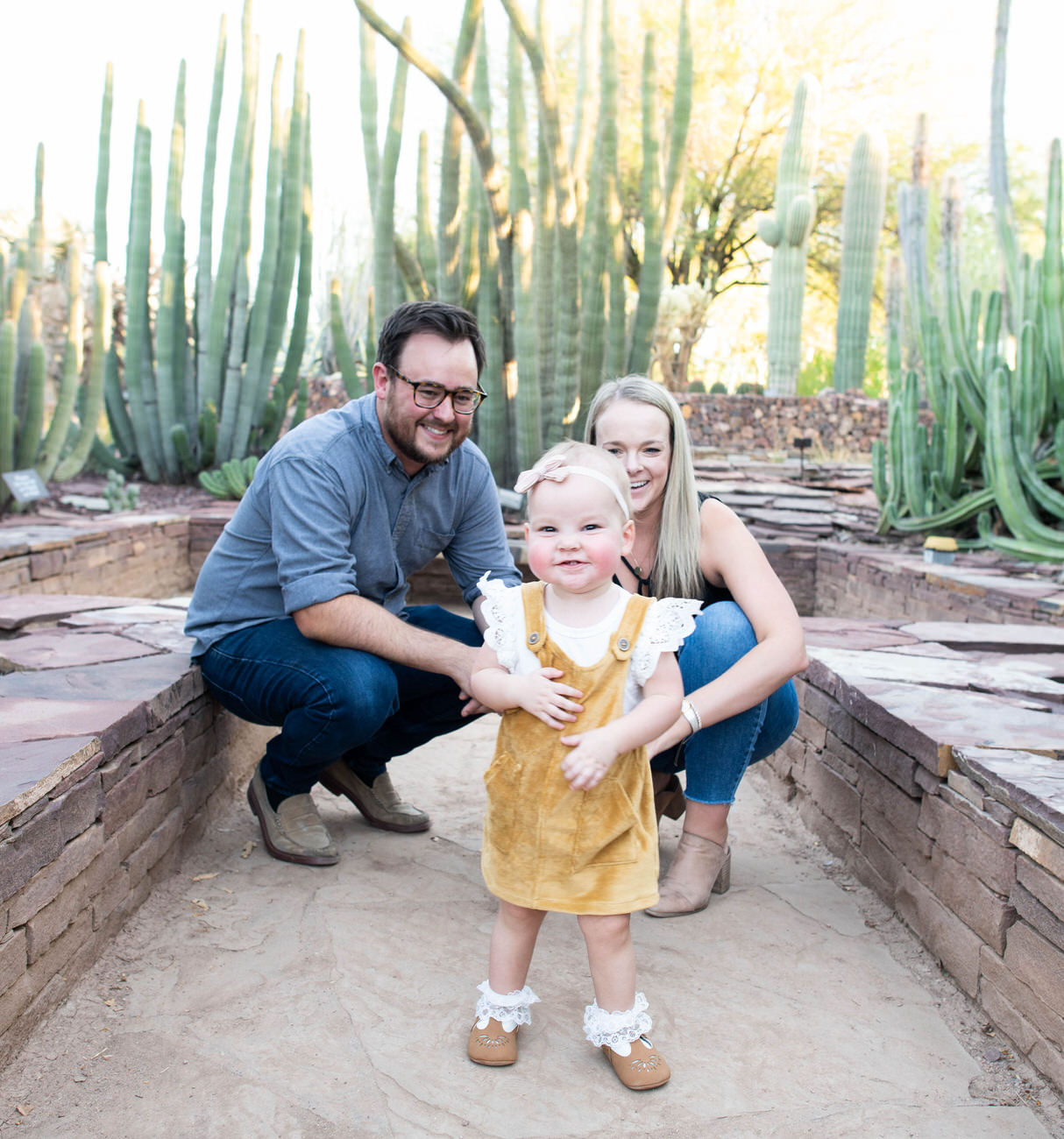 A family portrait with a man in a gray shirt, a woman in a black top, and their baby in the center, all smiling in front of a cactus backdrop at the Desert Botanical Garden in Arizona
