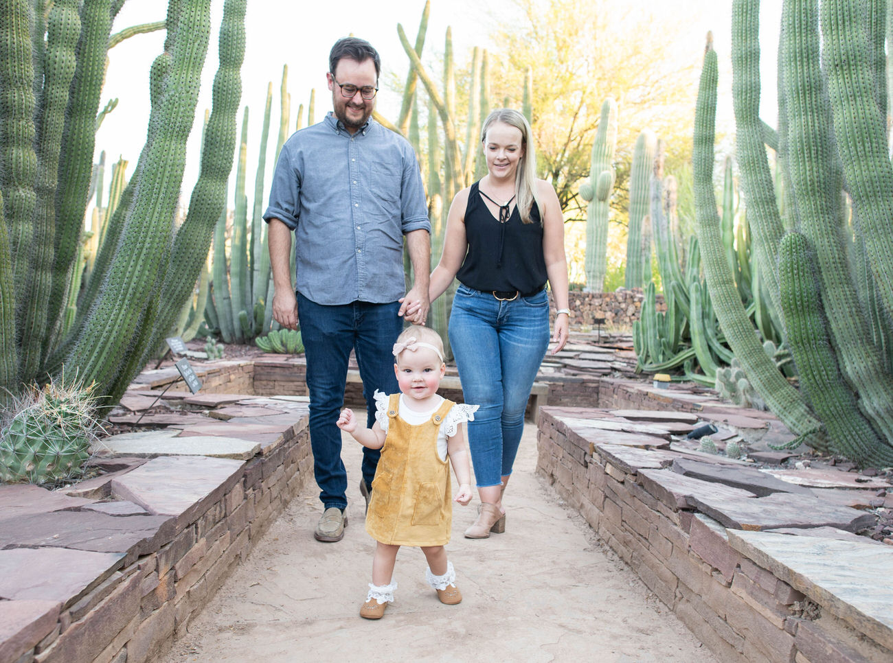 A family of three walking hand-in-hand through the Desert Botanical Garden in Arizona, with a toddler in a yellow dress leading the way amid a cactus-lined path.