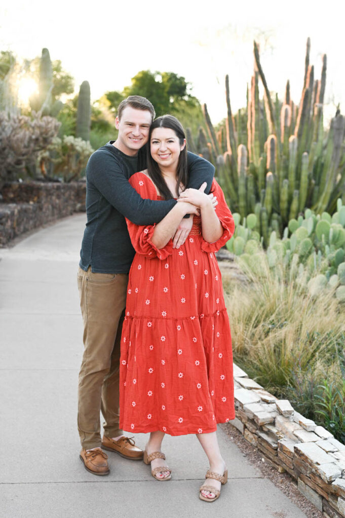 A smiling couple embracing, with the woman wearing a red patterned dress, in front of a desert landscape with tall cacti at the Desert Botanical Garden in Arizona, during sunset