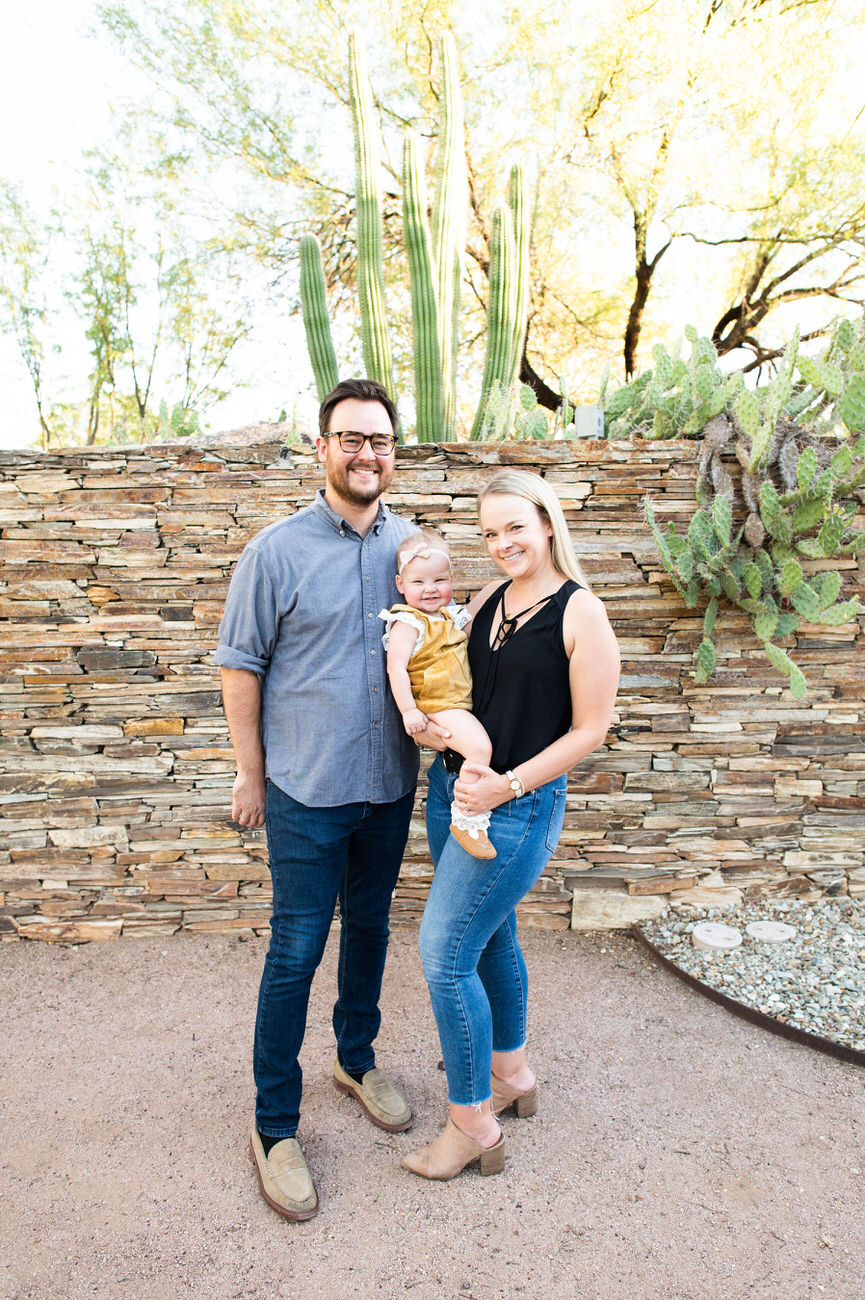 A family of three, with a man in a gray shirt, a woman in a black tank top, and a baby in a yellow dress, smiling in front of a cactus garden wall at the Desert Botanical Garden in Arizona.