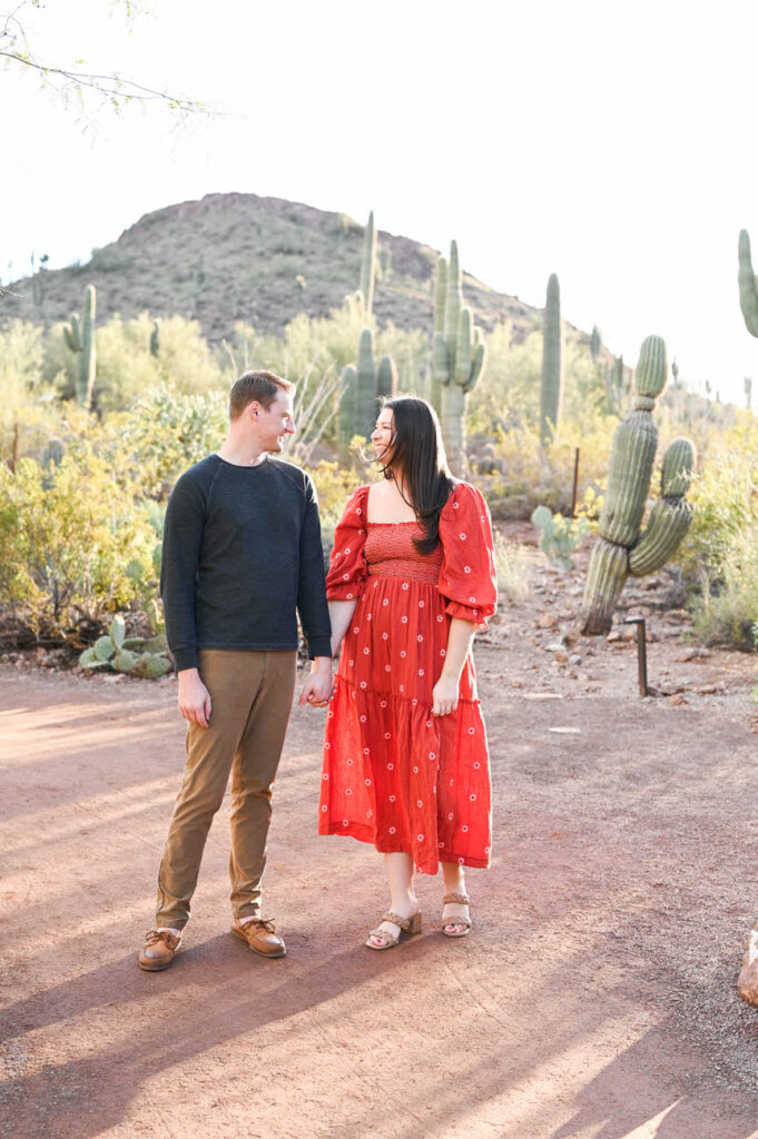 A couple holding hands and gazing at each other, with the woman in a red patterned dress, amidst a desert pathway lined with cacti at the Desert Botanical Garden in Arizona