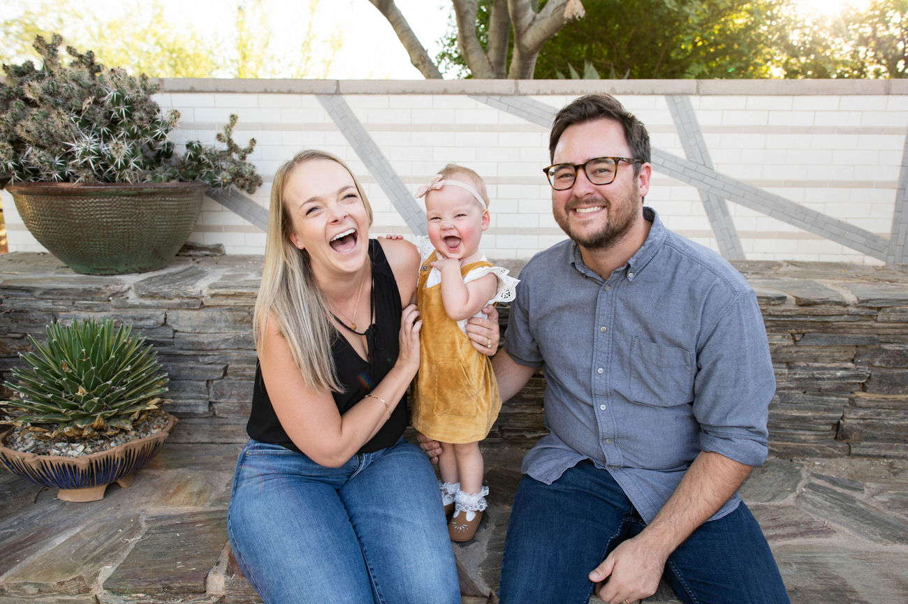 A joyous family moment with a laughing couple and their baby girl in front of a textured garden wall, surrounded by desert plants at the Desert Botanical Garden in Arizona