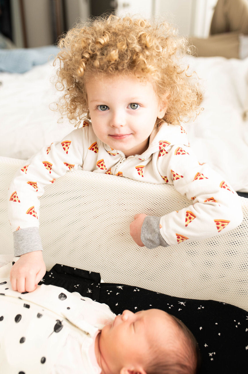 A toddler with curly blonde hair wearing a pizza slice patterned pajama smiles gently, peering over a mesh crib railing at a sleeping newborn.