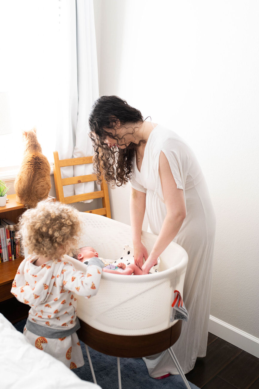 A curly-haired toddler and a ginger cat curiously watch a woman in a white dress leaning over a bassinet to comfort a newborn.