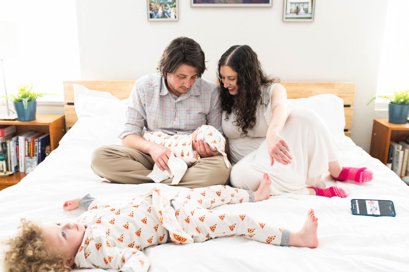 A casual family moment on a bed, with a man in a plaid shirt and a woman in a white dress attending to a baby, while a toddler in pizza slice patterned pajamas lies beside them.