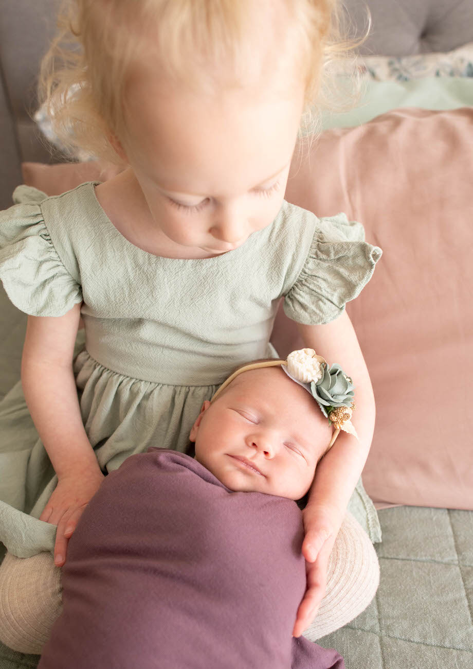 A top-down view of a blonde toddler in a green dress tenderly holding a sleeping newborn wrapped in a purple cloth.