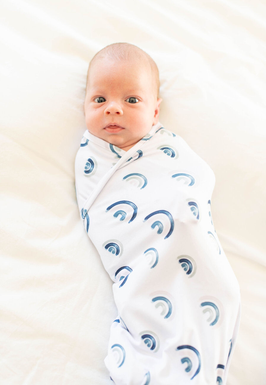 A newborn baby swaddled in a white cloth with blue rainbows lies on a bed, gazing upwards with wide eyes.
