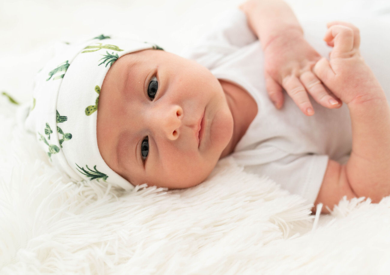 A newborn baby wearing a cactus-print headband lies on a fluffy white rug, looking curiously at the camera.