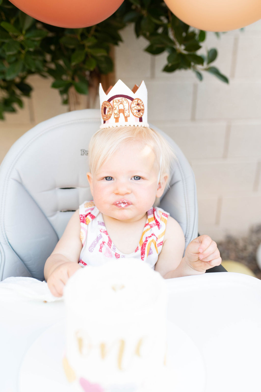 one year old blonde girl with a one year crown with a cake on her high chair tray looking at the camera